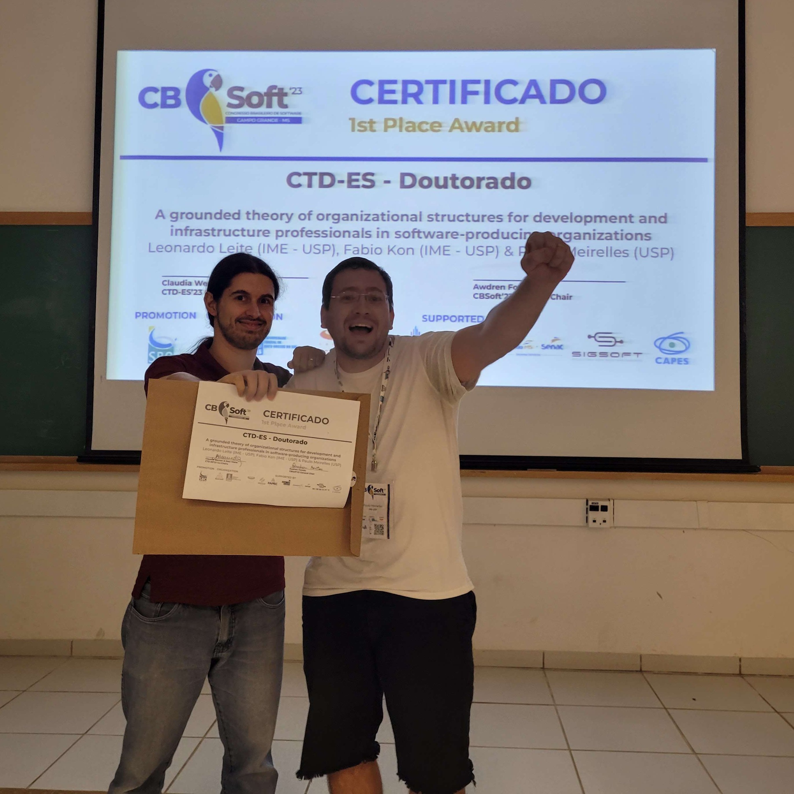Leonardo and Paulo holding the award certificate; Paulo celebrating with raised fist; behind a big screen displaying the award certificate.