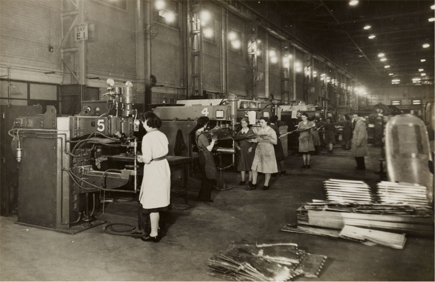 Operadoras mulheres em uma fábrica operando máquinas grandes, um homem supervisionando, foto preto e branco, pessoas vestindo trajes da década de 40 ou 50.